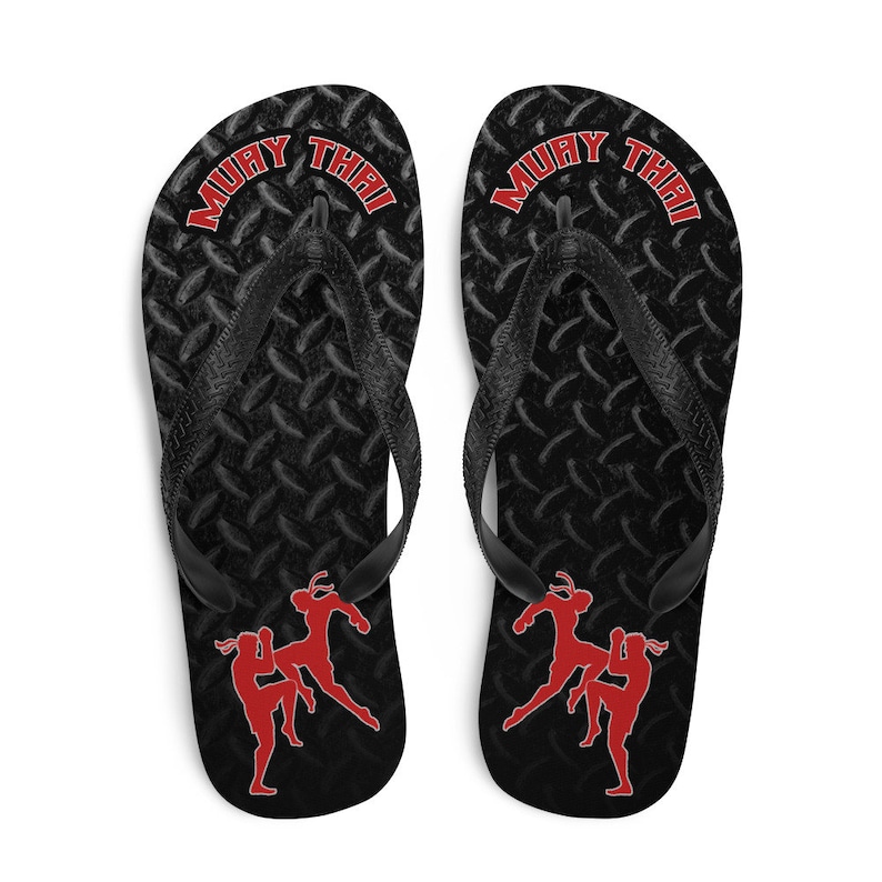 May include: Pair of black flip flops with a black and grey diamond pattern. The flip flops have the words "Muay Thai" in red on the straps and a red silhouette of two women practicing Muay Thai.