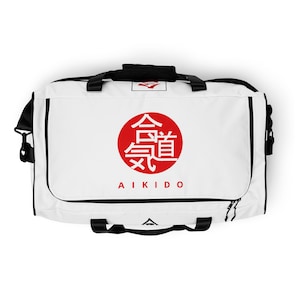 May include: White duffel bag with black trim and handles. A red circle with Japanese characters and the word "AIKIDO" in black lettering is centered on the front. The bag has a top handle and a shoulder strap.