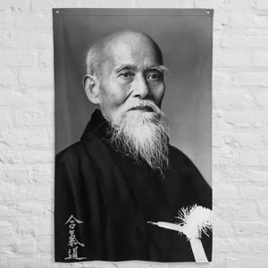 以下が含まれることがあります： 長い白いひげと伝統的な日本の衣装を着た男性の肖像画が描かれた、白黒の壁掛け旗。旗の左下には合気道の漢字が書かれています。