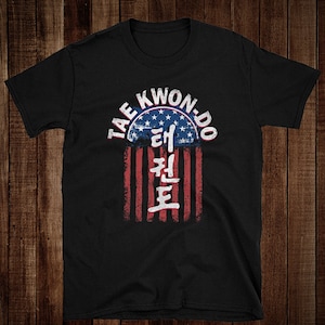 Puede incluir: Camiseta negra con un diseño de bandera estadounidense desgastado y el texto "Tae Kwon-Do" en un círculo sobre la bandera. El texto "태권도" está escrito en coreano debajo de la bandera.