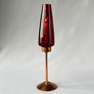 Könnte beinhalten: Ein hoher, roter Glas-Kerzenhalter mit einem kupfernen Sockel und Stiel. Der Kerzenhalter ist für eine einzelne Kerze ausgelegt.