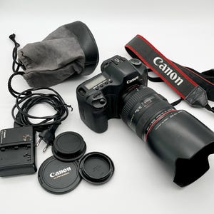 Op de afbeelding: Zwarte Canon DSLR-camera met lens, zonnekap en riem. Accessoires zijn onder meer een oplader, lensdoppen en een grijze draagtas. De riem is rood en zwart met het Canon-logo.