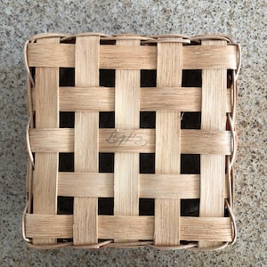 May include: A square woven basket lid made of light tan wooden splints arranged in a grid pattern. The center of the lid features a handwritten inscription that reads BGP 20 in black ink, integrated into the woven structure of the natural wood.