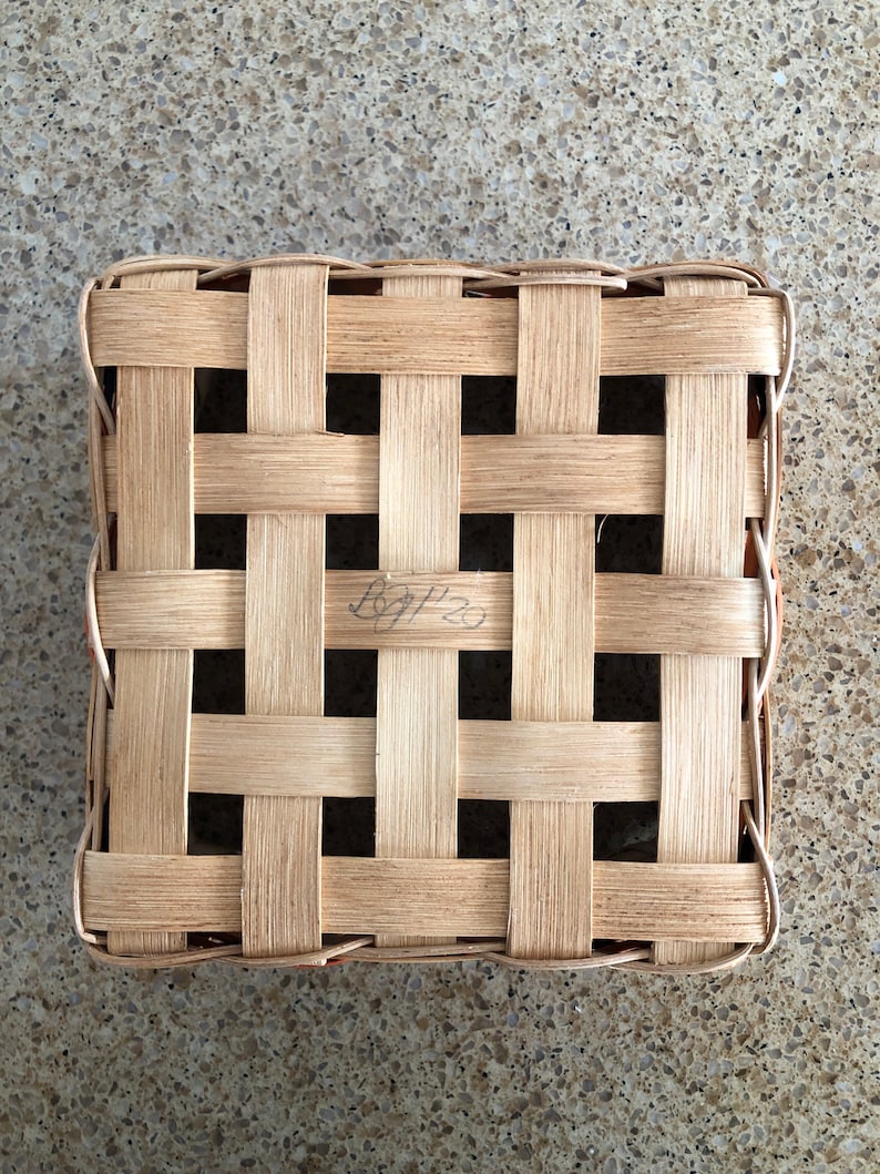 May include: A square woven basket lid made of light tan wooden splints arranged in a grid pattern. The center of the lid features a handwritten inscription that reads BGP 20 in black ink, integrated into the woven structure of the natural wood.
