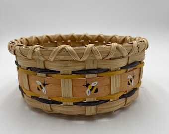 May include: A round woven basket with a brown and yellow striped band. The band is decorated with three painted bumblebees.