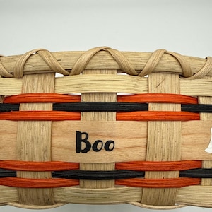 May include: A woven basket featuring light tan natural wood splints with horizontal bands of orange and black dyed reed. The center of the basket displays the word Boo in black lettering, integrated into the woven structure of the basket.