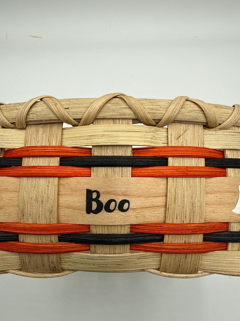 May include: A woven basket featuring light tan natural wood splints with horizontal bands of orange and black dyed reed. The center of the basket displays the word Boo in black lettering, integrated into the woven structure of the basket.