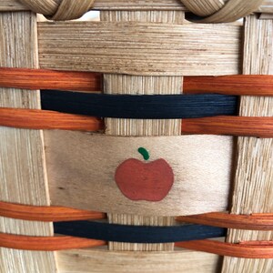 May include: A woven basket featuring a light tan natural wood finish with horizontal accents in black and orange. A small, painted red apple with a green stem is centered on a wide, flat wooden splint that forms part of the basket's structure.