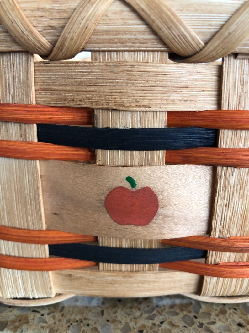 May include: A woven basket featuring a light tan natural wood finish with horizontal accents in black and orange. A small, painted red apple with a green stem is centered on a wide, flat wooden splint that forms part of the basket's structure.