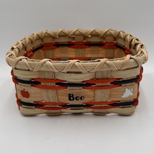 May include: A rectangular woven basket made of light tan wood splints with horizontal orange and black accent bands. The front features the word Boo in black lettering, a small red apple illustration, and a white ghost figure decoration.