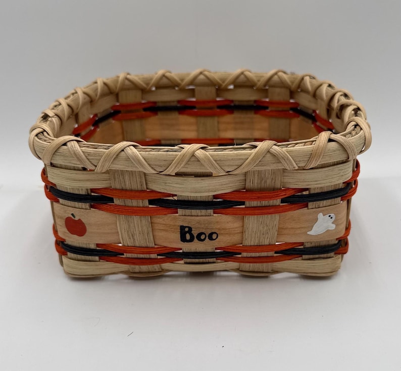 May include: A rectangular woven basket made of light tan wood splints with horizontal orange and black accent bands. The front features the word Boo in black lettering, a small red apple illustration, and a white ghost figure decoration.