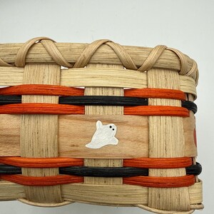 May include: A woven basket featuring natural light-colored reed strips interlaced with horizontal bands of vibrant orange and black dyed material. A small, hand-painted white ghost with two black dot eyes is centered on a smooth wooden slat.