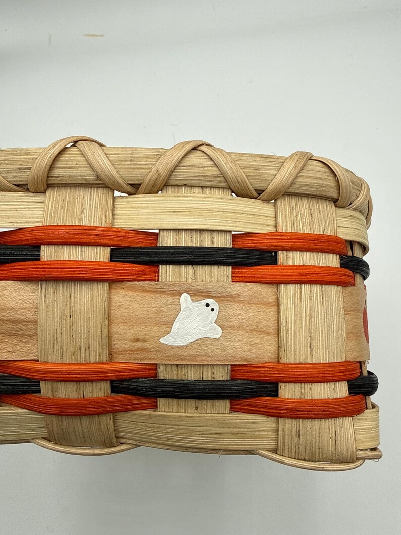 May include: A woven basket featuring natural light-colored reed strips interlaced with horizontal bands of vibrant orange and black dyed material. A small, hand-painted white ghost with two black dot eyes is centered on a smooth wooden slat.