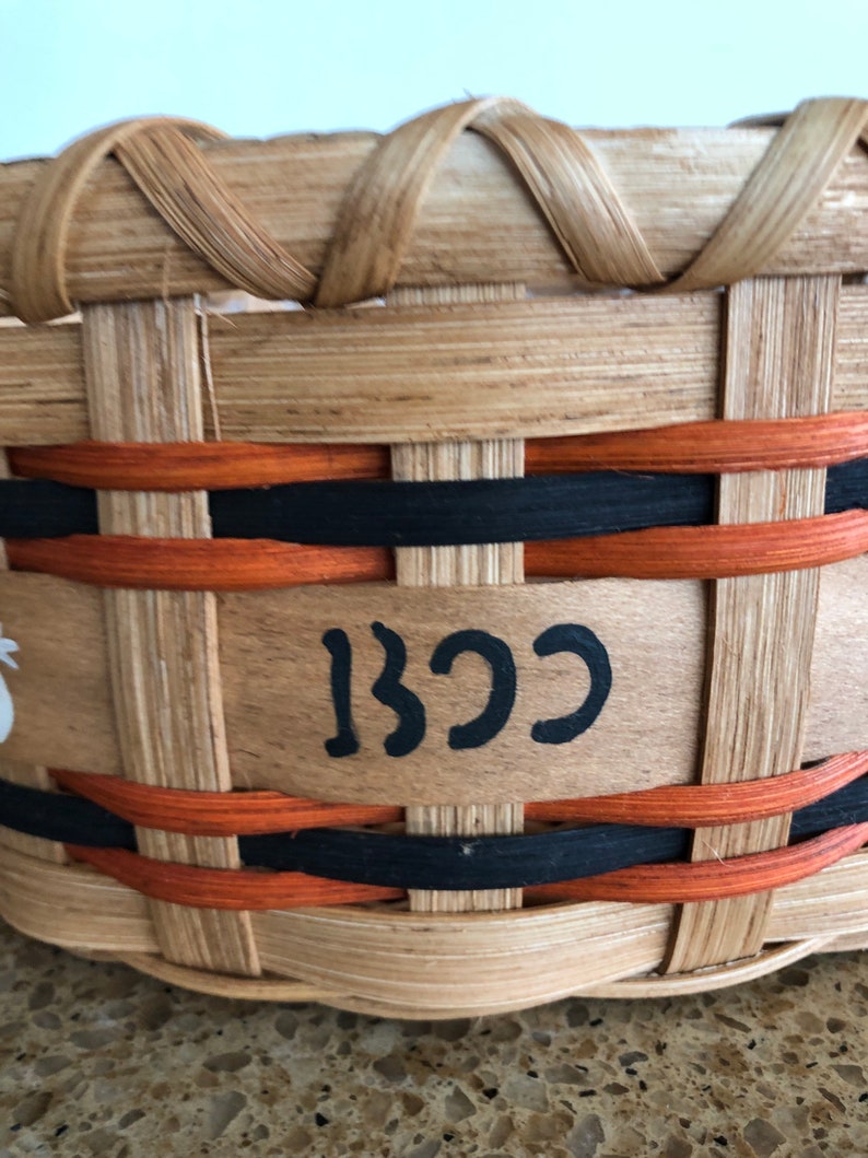 May include: A woven basket with black and orange trim. The basket has a wooden base with the word "BCC" written in black paint.
