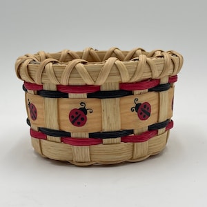 May include: A small, round woven basket with a red, black, and natural straw design. The basket has a band of red and black trim around the top and a band of red and black trim around the middle. The middle band has three ladybugs painted on it.
