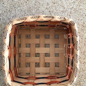 May include: A square woven basket featuring light tan wooden splints in a grid pattern. The upper rim is reinforced with a twisted rope border, while the interior sides are accented with thin, horizontal orange and black decorative strips.