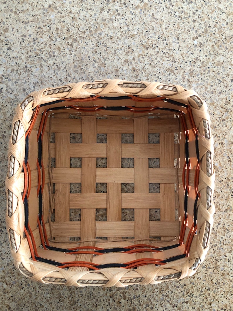 May include: A square woven basket featuring light tan wooden splints in a grid pattern. The upper rim is reinforced with a twisted rope border, while the interior sides are accented with thin, horizontal orange and black decorative strips.