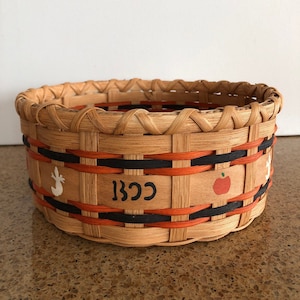 May include: A woven basket with a natural wood finish and black and orange trim. The basket has a white painted design of a deer and an apple on the front. The text "BCC" is painted on the front of the basket.