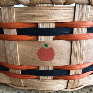 May include: A woven basket with black and orange trim and a painted apple design. The basket is made of natural wood.