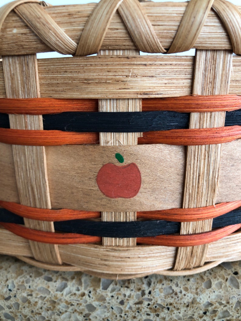 May include: A woven basket with black and orange trim and a painted apple design. The basket is made of natural wood.