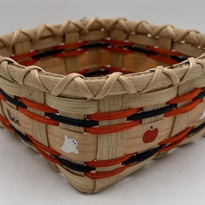 May include: A square woven basket featuring light tan natural reeds accented with horizontal orange and black bands. The exterior displays painted white ghost figures, a red apple, and the hand-painted black text "Boo" on the side.