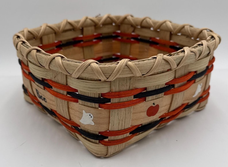 May include: A square woven basket featuring light tan natural reeds accented with horizontal orange and black bands. The exterior displays painted white ghost figures, a red apple, and the hand-painted black text "Boo" on the side.