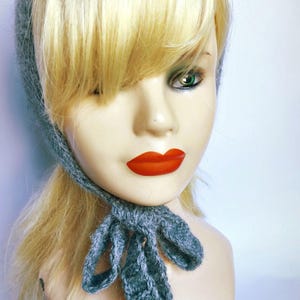May include: A gray knitted hat with a matching tie detail. The hat is worn by a mannequin with blonde hair, red lipstick, and green eyes. The hat is a soft, textured material.