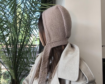 May include: A beige knitted bonnet with a textured stitch pattern and long, flat knitted ties that hang down from the sides. The bonnet features a structured seam along the top and back, providing a close-fitting shape that covers the head and ears.