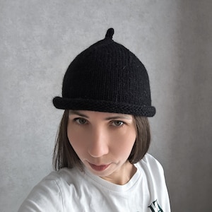 Może przedstawiać: Czarny dzianinowy kapelusz z zaokrągloną koroną i małym rondem. Kapelusz ma mały, zaokrąglony detal na górze. Osoba nosi białą koszulkę z zielonym tekstem.