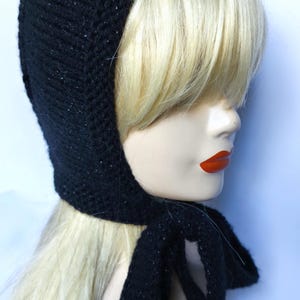 May include: A black knitted hood with a tie closure, displayed on a mannequin head with blonde hair. The hood is made of a dark, textured material with subtle flecks of sparkle.