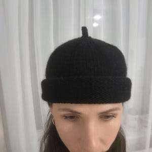 May include: A solid black knit beanie cap featuring a rounded crown with a small, centered top knot detail and a thick, folded brim. The hat is constructed from a textured yarn material, providing a consistent dark appearance throughout the design.