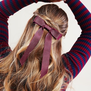 May include: A woman with long, wavy, light brown hair with a burgundy ribbon tied in a bow in her hair.