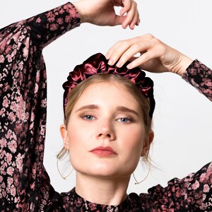 May include: A woman wearing a black and pink patterned dress with a burgundy velvet headband. She is posing with her arms raised above her head.
