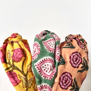 May include: Three fabric headbands with floral patterns. One is yellow with pink flowers, another is green with pink and white paisley shapes, and the third is peach with pink roses. The headbands are bunched together.