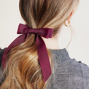 Puede incluir: Una cinta para el pelo con lazo de color burdeos atada en una coleta. La cinta para el pelo está hecha de un material satinado y tiene un diseño simple y elegante.