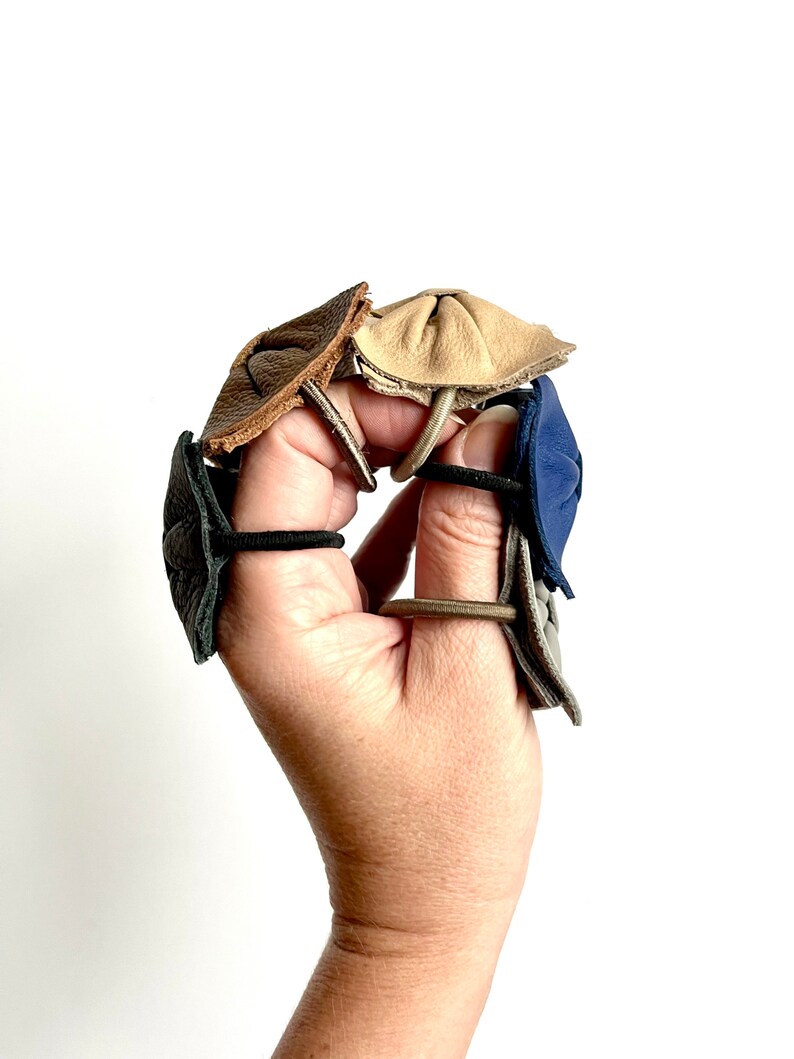 May include: Four leather hair tie holders in different colors: brown, beige, gray, and blue. The holders are made of leather and have a unique design.