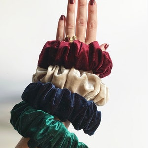 May include: A set of four scrunchies in different colors: burgundy, beige, navy blue, and green. The scrunchies are made of velvet and have a gathered, ruffled texture.