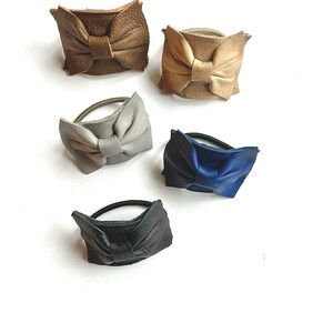 May include: Five leather hair ties with bow accents in different colors: brown, beige, gray, blue, and black.