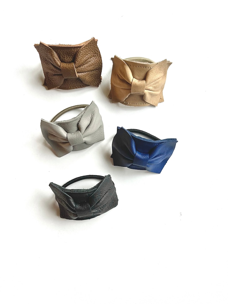 May include: Five leather hair ties with bow accents in different colors: brown, beige, gray, blue, and black.