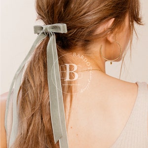 Puede incluir: Una mujer con el pelo largo y castaño atado con un lazo de terciopelo verde salvia. La cinta está atada en un nudo simple y los extremos se dejan largos. La mujer lleva un top de tirantes beige claro y pendientes de aro dorados.