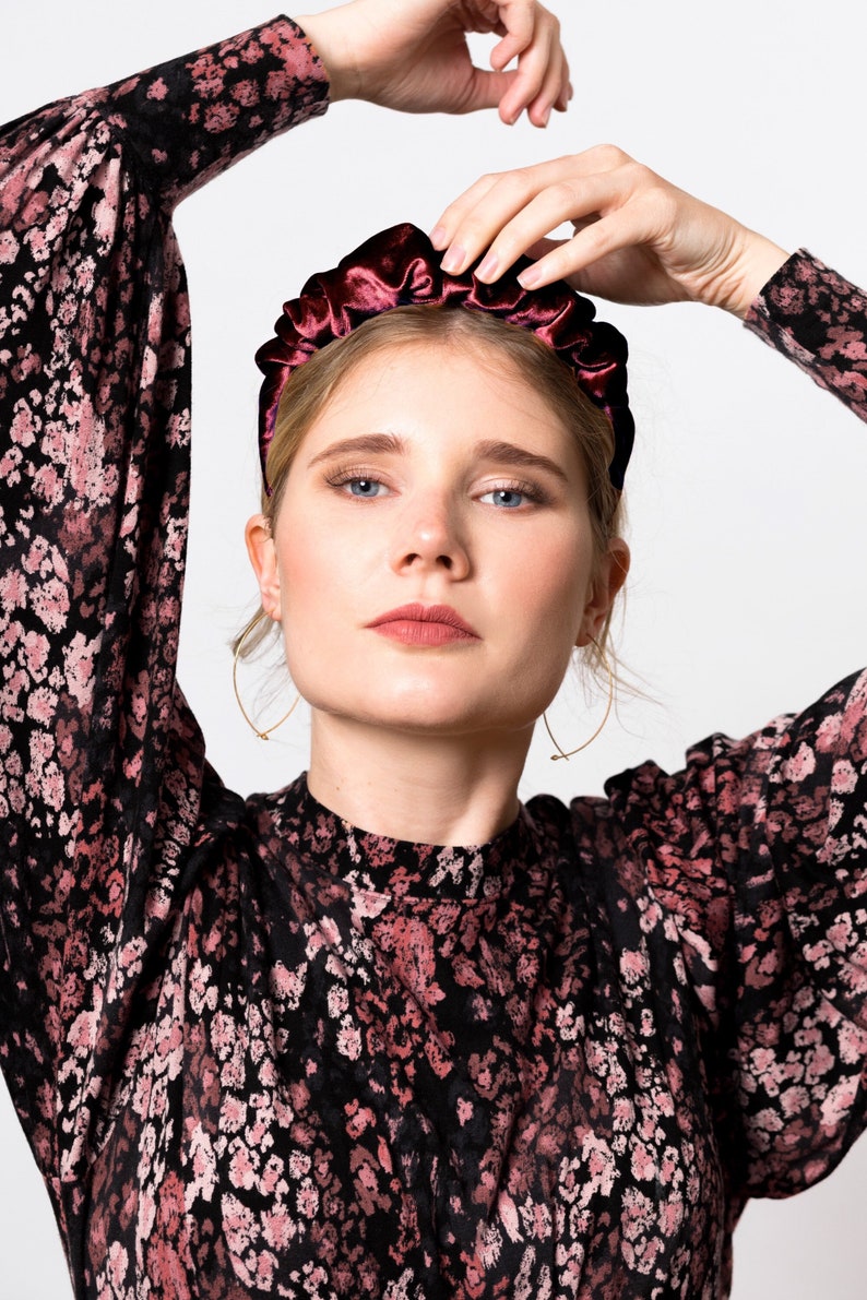 May include: A woman wearing a black and pink patterned dress with a burgundy velvet headband. She is posing with her arms raised above her head.