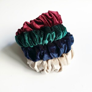 May include: Three velvet scrunchies in burgundy, dark green, and navy blue colors. The scrunchies are stacked on top of each other.
