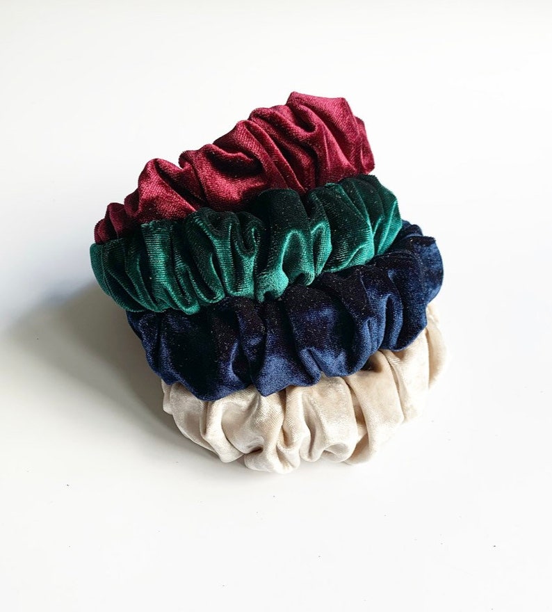 May include: Three velvet scrunchies in burgundy, dark green, and navy blue colors. The scrunchies are stacked on top of each other.
