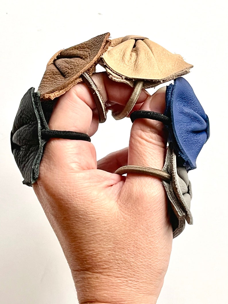 May include: Five leather fingertip protectors in different colors: black, brown, beige, gray, and blue. Each protector is attached to a hair elastic.