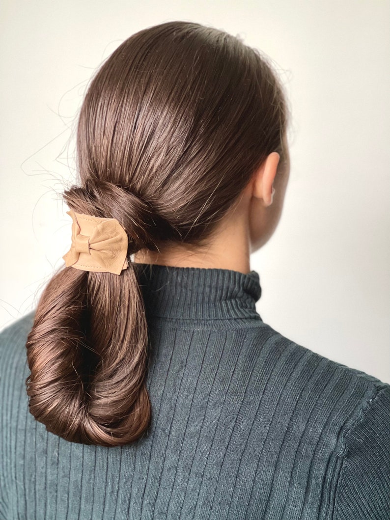 May include: A woman with long brown hair tied back in a low ponytail with a brown leather hair tie.