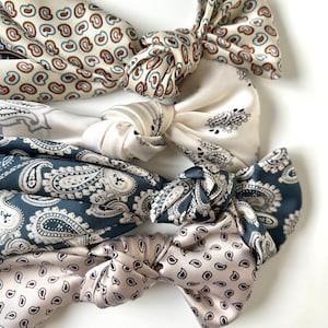 Könnte beinhalten: Vier Seiden-Haarbänder in verschiedenen Farben und Mustern. Die Haarbänder sind in der Mitte geknotet und zeigen Paisley- und andere dekorative Designs in Creme, Marineblau und Mauve. Die Haarbänder sind aus einem glänzenden, seidigen Material gefertigt.
