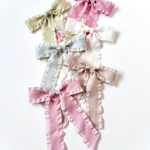 May include: A set of six ruffled ribbon hair bows in various pastel colors, including pink, green, and beige. The bows are arranged in a pile, with the pink bow on top.