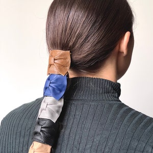 May include: A ponytail holder with four leather bows in brown, blue, gray, and beige.