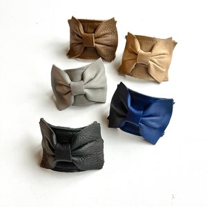 May include: Five leather bow hair clips in different colors: brown, beige, gray, blue, and black.
