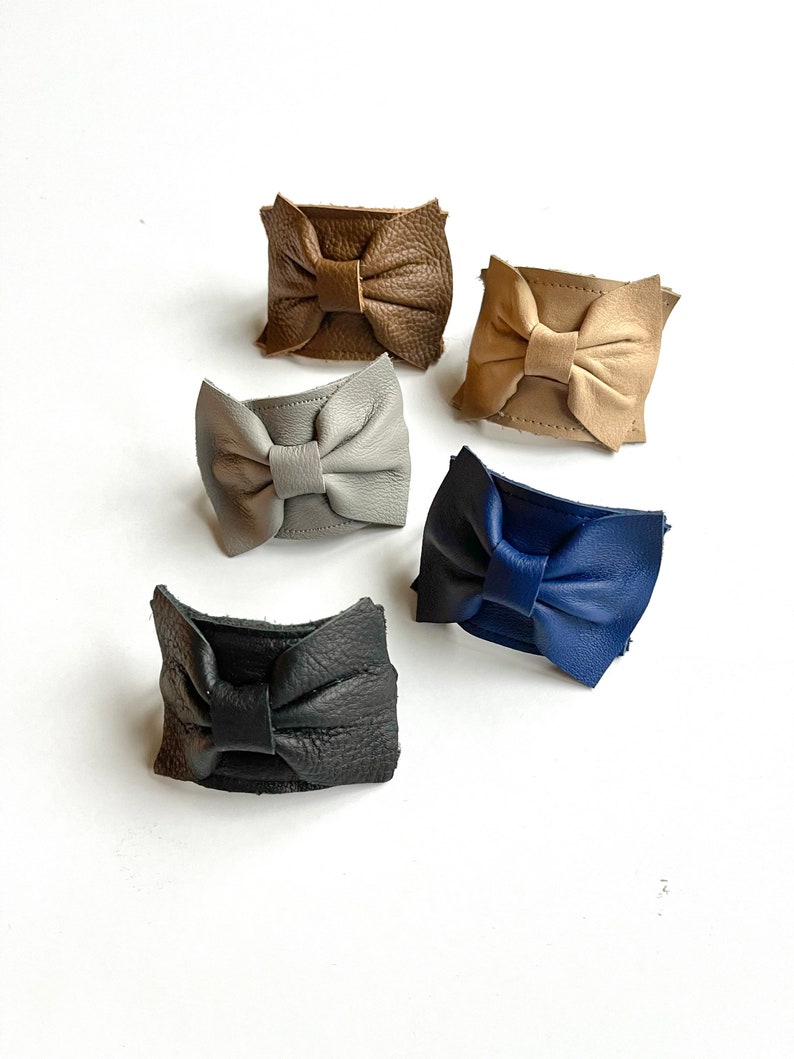 May include: Five leather bow hair clips in different colors: brown, beige, gray, blue, and black.